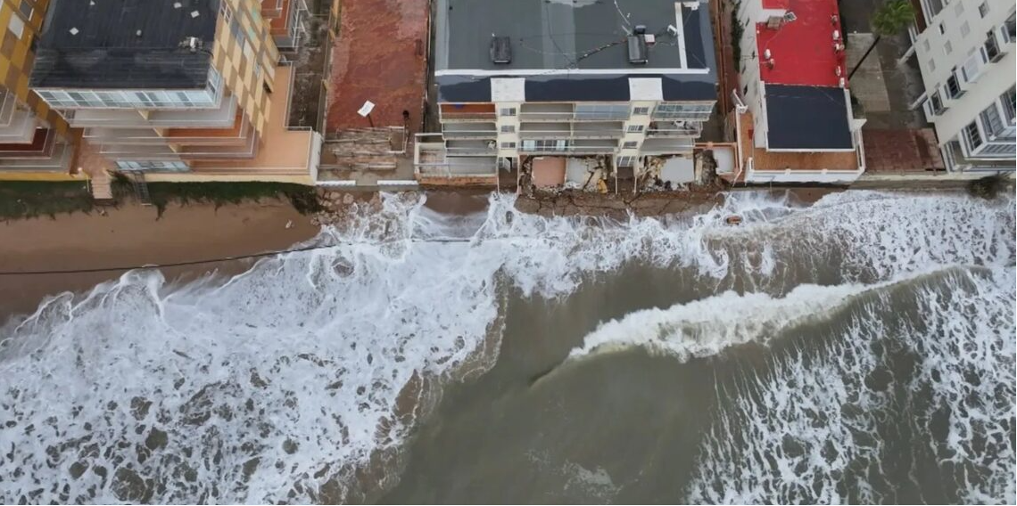 La indignació veïnal i la preocupació pel futur de la costa han tornat a prendre el passeig marítim de Tavernes de la Valldigna en una nova jornada de protestes. Centenars de residents s'han mobilitzat per a exigir a la Direcció General de Costes la regeneració urgent del seu litoral, greument afectat per la pèrdua d'arena i l'amenaça directa del mar sobre els habitatges. TAVERNES DE LA VALLDIGNA. 23 d'agost de 2026.— El malestar veïnal i la preocupació mediambiental han tornat a prendre els carrers de Tavernes de la Valldigna. Centenars de residents s'han mobilitzat aquest cap de setmana per a alçar la veu davant el que consideren una situació insostenible: la paulatina i imparable desaparició del seu front litoral a causa dels successius temporals marítims que afecten la zona. La pèrdua de superfície de platja ha aconseguit nivells sense precedents en el municipi. Segons els últims informes tècnics i mesuraments locals, algunes seccions de la costa han perdut fins a un 77% de la seua massa d'arena original, deixant el ferm de les zones habitades exposat a l'embat directe de l'onatge. Habitatges en perill i desallotjaments preventius La gravetat de l'erosió ha deixat de ser una mera pèrdua d'atractiu turístic per a convertir-se en un problema de seguretat pública. Els últims episodis de marejada i fort onatge van provocar l'esfondrament de murets de protecció i la infiltració d'aigua en les infraestructures costaneres, una situació que va obligar les autoritats locals a evacuar d'emergència diversos habitatges situats en primera línia de mar. Els propietaris afectats denuncien que la falta d'arena ha après el matalàs natural que abans amortia la força de les ones, deixant les fonamentacions directament exposades a la violència de la mar. Exigències a les administracions i silenci municipal La marxa pacífica va recórrer el passeig marítim exigint solucions definitives a la Direcció General de Costes i al Ministeri per a la Transició Ecològica. Els col·lectius veïnals reclamen superar la fase dels "parches d'emergència" —basats en l'aportació puntual d'arena que els temporals tornen a arrossegar— i executar projectes d'enginyeria de regeneració litoral duradors, com ara la instal·lació d'espigons o barreres submarines. Per la seua banda, el govern local ha evitat fer cap mena de publicitat de la convocatòria. Aquesta actitud manté la línia de l'alcaldessa, de signe socialista, qui igual que va evitar enfrontar-se amb el ministre Óscar Puente per a exigir la represa de les obres de l'estació d'ADIF, evita ara enfrontar-se amb els seus companys de partit a la Direcció General de Costes i a la Delegació del Govern. Els veïns, mentrestant, temen que l'arribada dels temporals de tardor i hivern cause danys estructurals irreparables a la localitat.